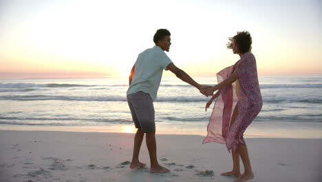 Una-Pareja-Birracial-Se-Toma-De-La-Mano-En-Una-Playa-Al-Atardecer,-Las-Olas-Rompiendo-Suavemente-En-El-Fondo