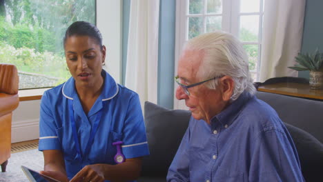 senior couple at home with man talking to female nurse or care worker using digital tablet
