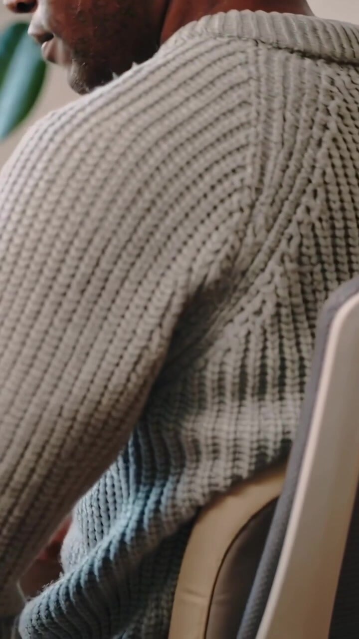 Video verticale: Uomo con maglione a coste che si allunga per sistemare lo schienale imbottito della sedia dopo aver provato dolore vicino a una pianta d'ufficio.