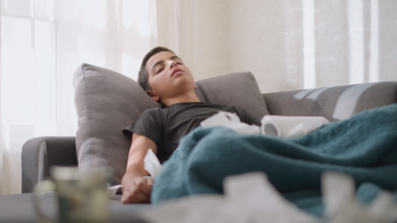 Close-up of sick boy lying down on couch, weak and drained of energy, surrounded by tissues, with light shining through window, he rests and struggles with fatigue from illness or flu symptoms