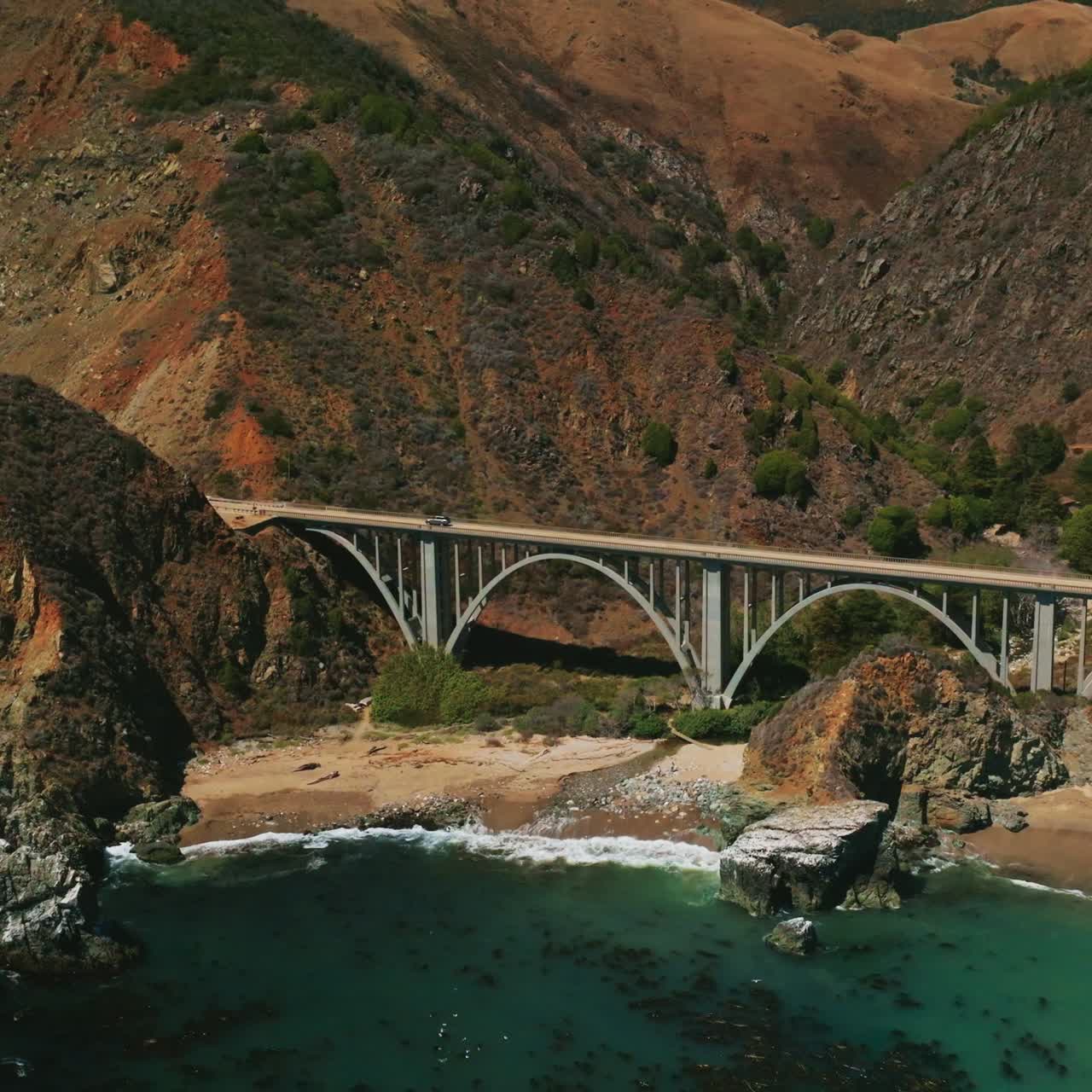 Arched bridge in the mountains of California with cars moving by. Drone footage rising over the pacific ocean shore on sunny daytime