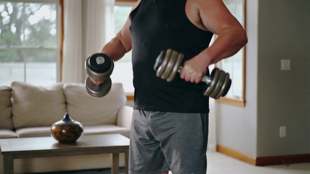 Beginning workout man in black tank, grey shorts lifting adjustable dumbbells on dark mat at home