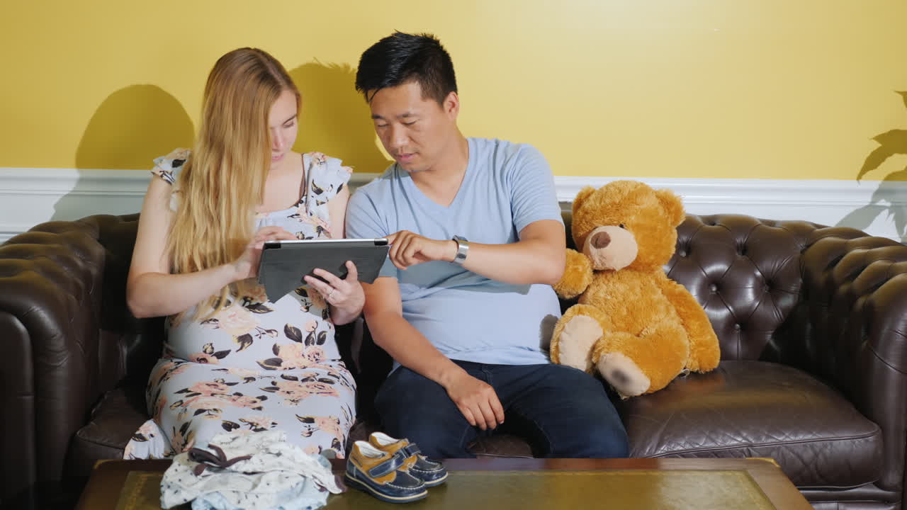 젊은 다민족 부부는 태어나지 않은 아기를 위해 옷을 주문하고 태블릿을 사용합니다.