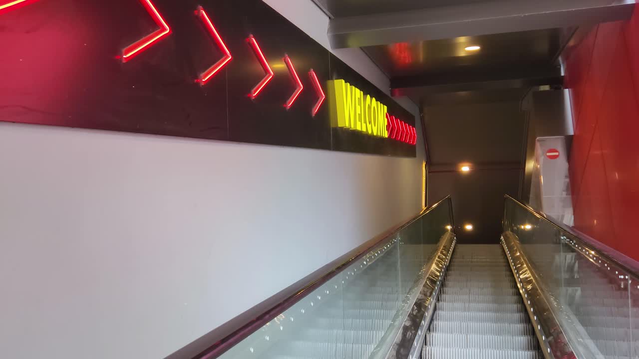 Cinematic upward-moving escalator leading to a glowing neon “WELCOME” sign with directional red arrows