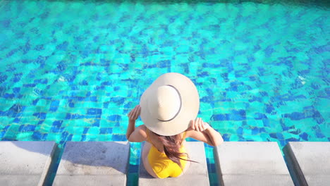 Vista-Superior-De-Una-Pequeña-Mujer-Con-Traje-De-Baño-Amarillo-Y-Sombrero-Flexible-Sentado-Junto-A-La-Piscina-En-Un-Destino-Tropical-En-Un-Día-Soleado,-Fotograma-Completo