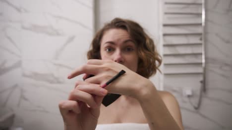 Young-woman-in-bath-towel-doing-eyebrow-makeup-in-bathroom---holding-a-penciland-explaining-to-camera-how-to-make-perfect-eyebrows.-Morning-make-up