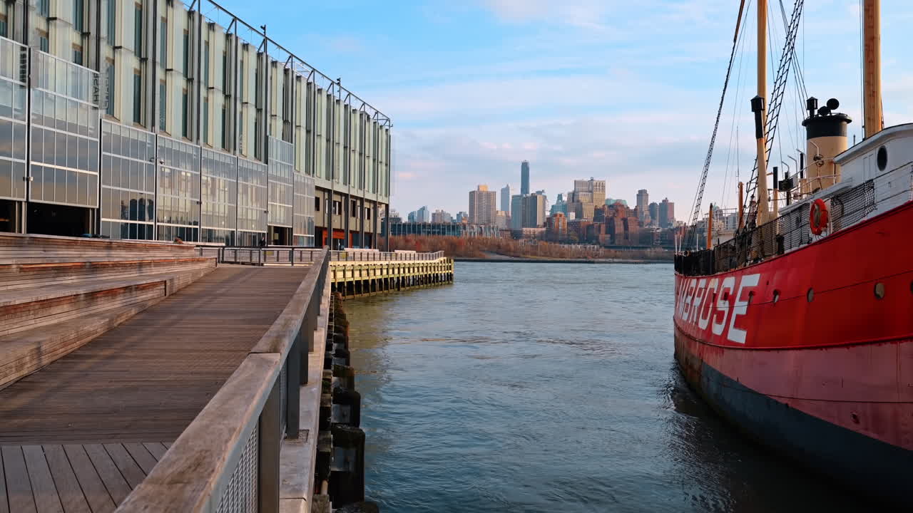 Molo e ponte sul lungomare con vista sullo skyline di Brooklyn. Una grande nave dipinta di rosso è ormeggiata a un molo accanto a un ponte di legno sul lungomare, con vista sullo skyline di Brooklyn in lontananza.