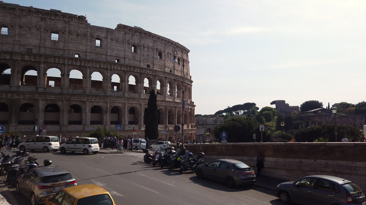 The colosseum from a different side than usual on a sunny spring day. There are few cars parked on the sides of the street. Cars are passing by. People are walking on the sidewalk. slow-motion.