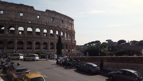 the colosseum from a different side than usual on a sunny spring day. there are few cars parked on the sides of the street. cars are passing by. people are walking on the sidewalk. slow-motion.