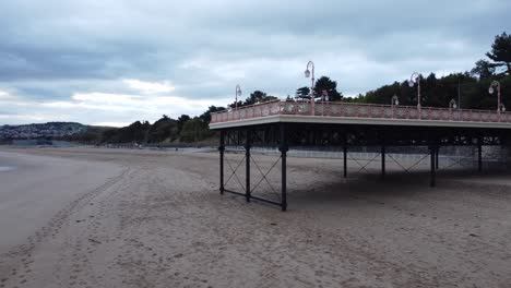 Colwyn-Bay-Welsh-seaside-town-pier-boardwalk-aerial-view-over-moody-overcast-sandy-low-tide-beach-close-low-orbit-left