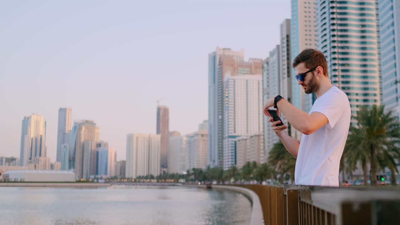 Happy handsome man in sunglasses and white t-shirt with a beard taking photos on a smartphone while standing on the waterfront in the summer town in the background and buildings