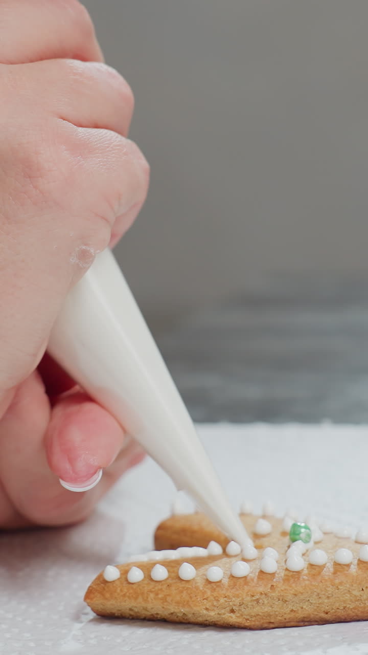 vista lateral de primer plano de la mano decorando una galleta en forma de amor con puntos blancos de glaseado y una perla verde, la galleta se coloca en papel de textura blanca mientras el glaseado se aplica cuidadosamente usando una bolsa de tubería