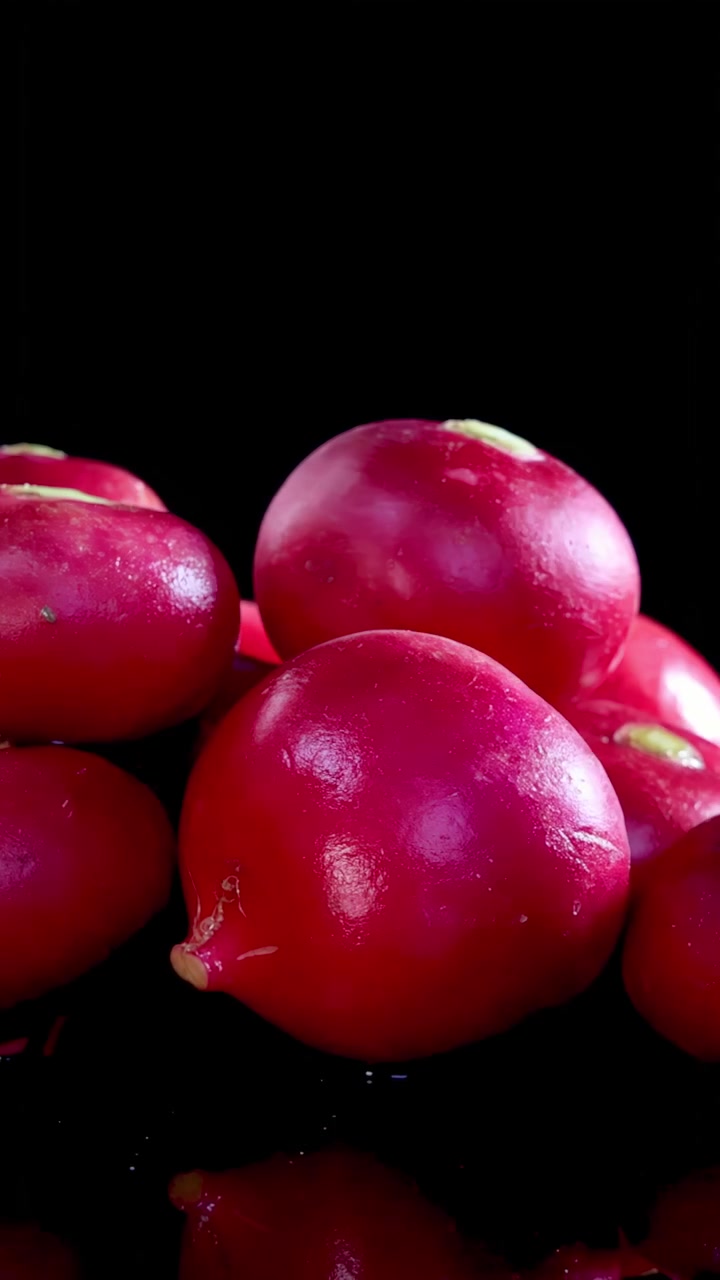 A cluster of glossy red radishes rotates smoothly on a reflective black surface, illuminated by dramatic studio lighting for a vibrant, high-contrast effect