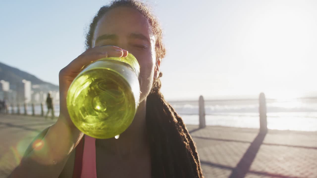mujer afroamericana en ropa deportiva bebiendo agua en el paseo marítimo
