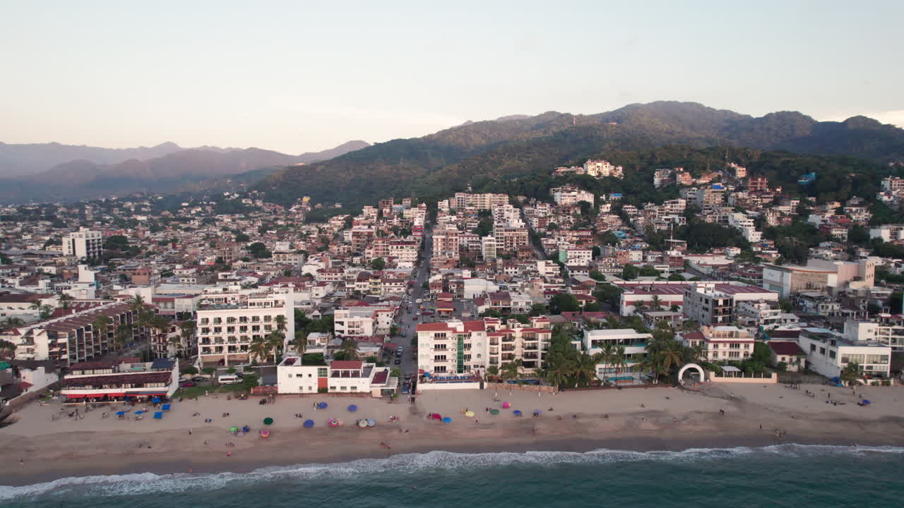 Video de dron mostrando una toma transversal de la costa de Puerto Vallarta al atardecer, destacando el mar, la playa, la ciudad y las montañas de la región.