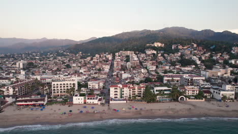 Drone-video-featuring-a-transverse-shot-of-the-coast-of-Puerto-Vallarta-at-sunset,-showcasing-the-sea,-beach,-city,-and-mountains-of-the-region