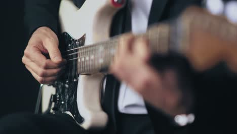 close up of a professional musician in a black suit playing chords on a white electric stratocaster guitar during a live session with a dark background