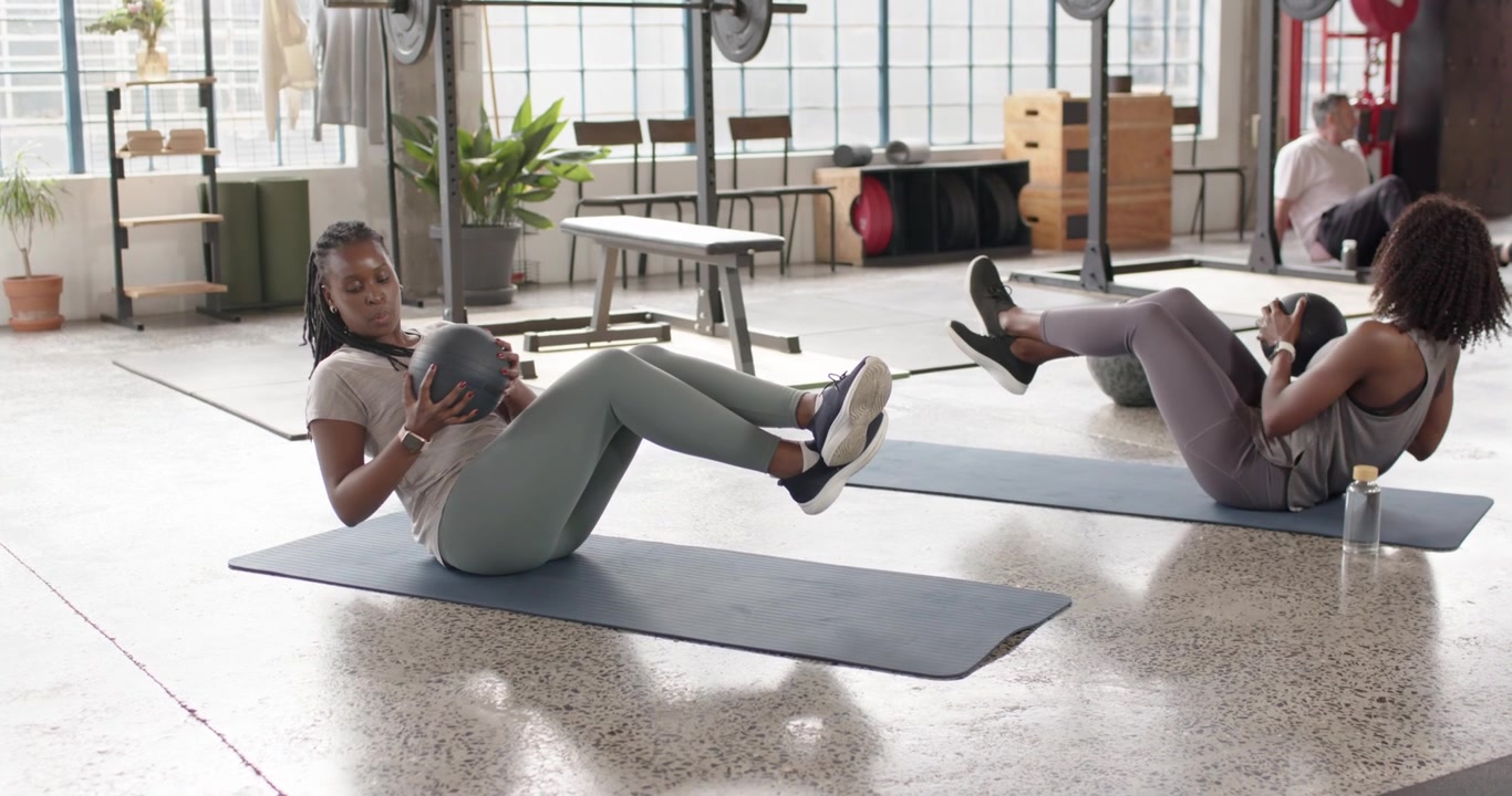 African American women starting tabletop on blue mats, twisting with medicine balls in gym for core