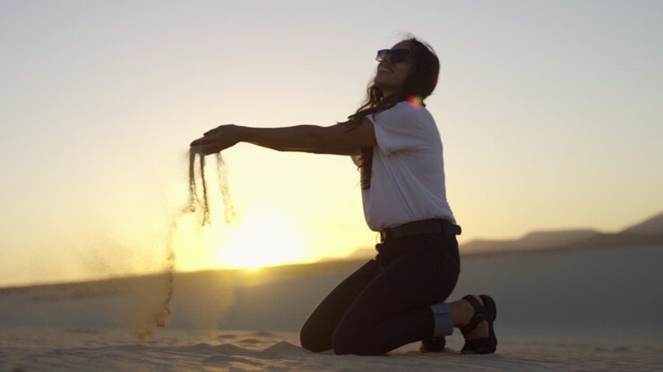 giovane donna latina in ginocchio dune di sabbia del deserto raccogliere una manciata di sabbia e versarla tra le dita contro il tramonto paesaggio naturale viaggio avventura in luoghi remoti inesplorati