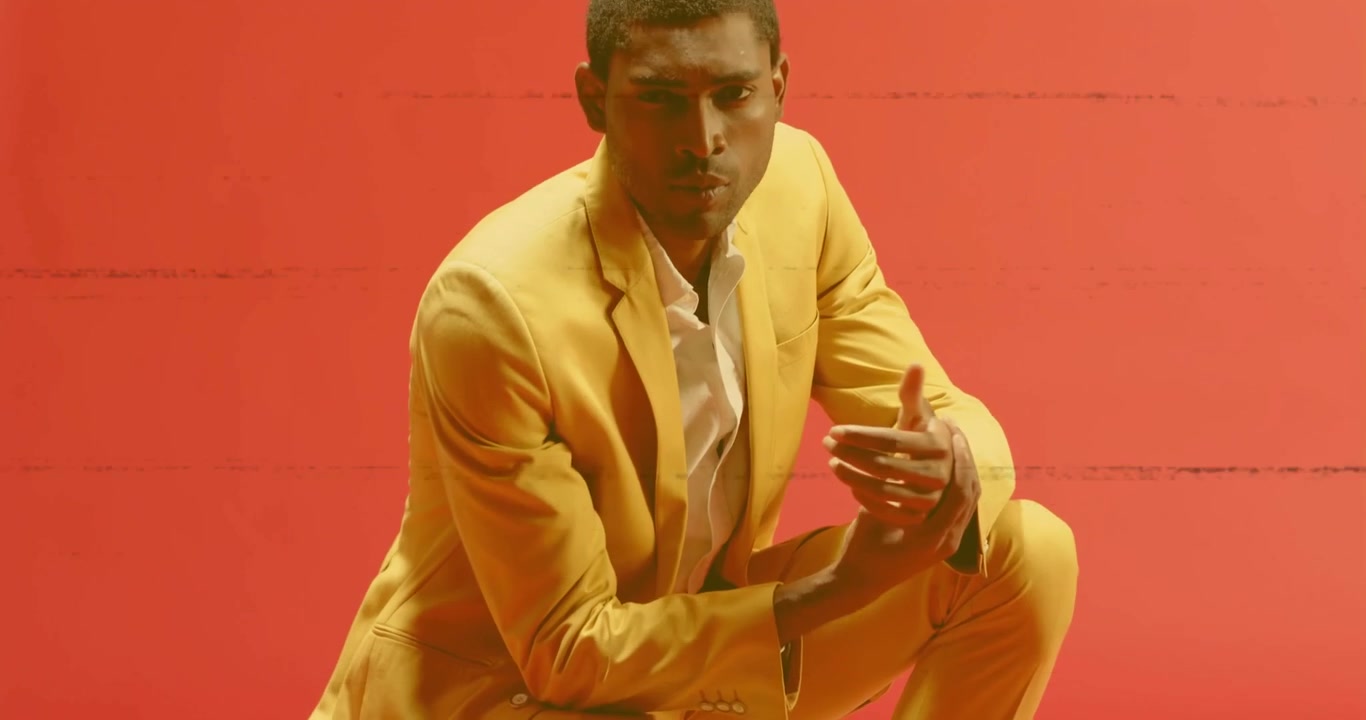 Camera panning back tilting upward showing mustard suit against red backdrop, man shifting on crate