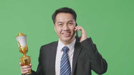 close up of asian business man in a suit and tie with a gold medal holding a gold trophy and talking on smartphone on green screen background in the studio