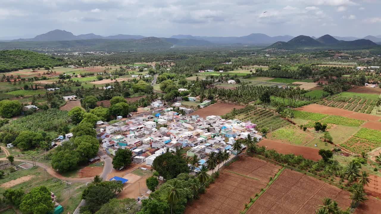 Cinematic footage contrasting urban sprawl with natural landscapes. The clip shifts from an established, rural village and its farmland to a nearby town with forested hills and mountains under a sky