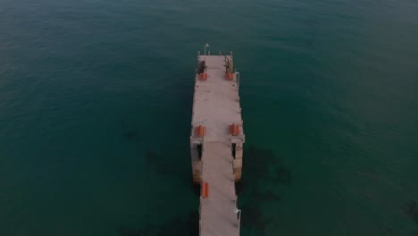 Backward-flight-above-green-calm-ocean-sea-waters-and-old-Porto-Santo-island-beach-pier-in-Portuguese-Madeiran-archipelago,-tilt-up-to-reveal-blue-sky-and-sand,-Portugal,-overhead-aerial-pull-back