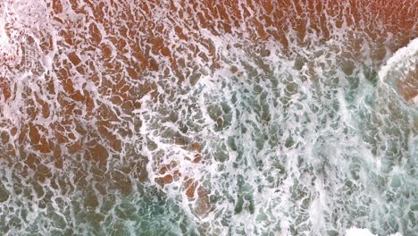 Vista-Aérea-De-Una-Ola-Rompiendo-En-Una-Playa-De-Arena-Dorada,-Los-Fascinantes-Patrones-De-Espuma-Y-Surf