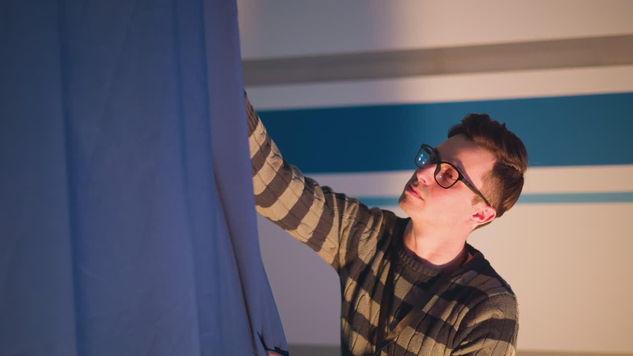 Male technician adjusts blue curtain on television studio set during behind scenes broadcast prep, floor spotlight illuminates striped backdrop while ladder, cables and rigging equipment