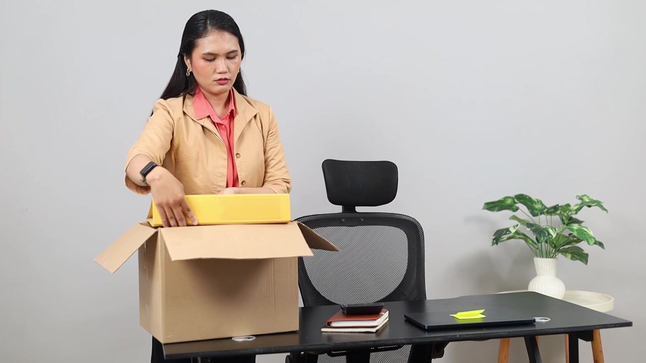 A female worker is in an office space tidying up things on her desk, putting them into a brown cardboard box, with a sad expression, the concept of layoffs or resignation.