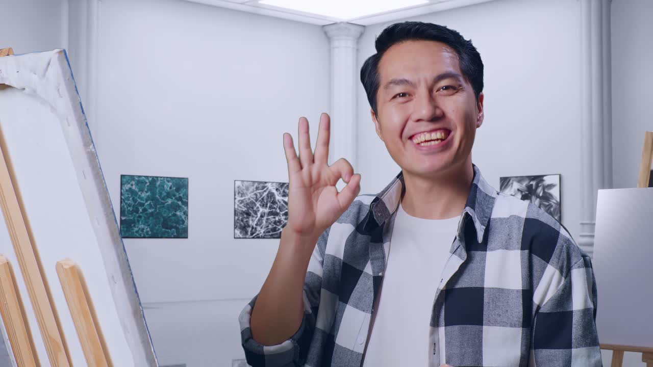 갤러리에서 오일 페인트와 으로 캔버스에 그림을 그리는 동안 미소 짓고 카메라에 은 몸짓을 보여주는 아시아 남성 예술가의 클로즈업