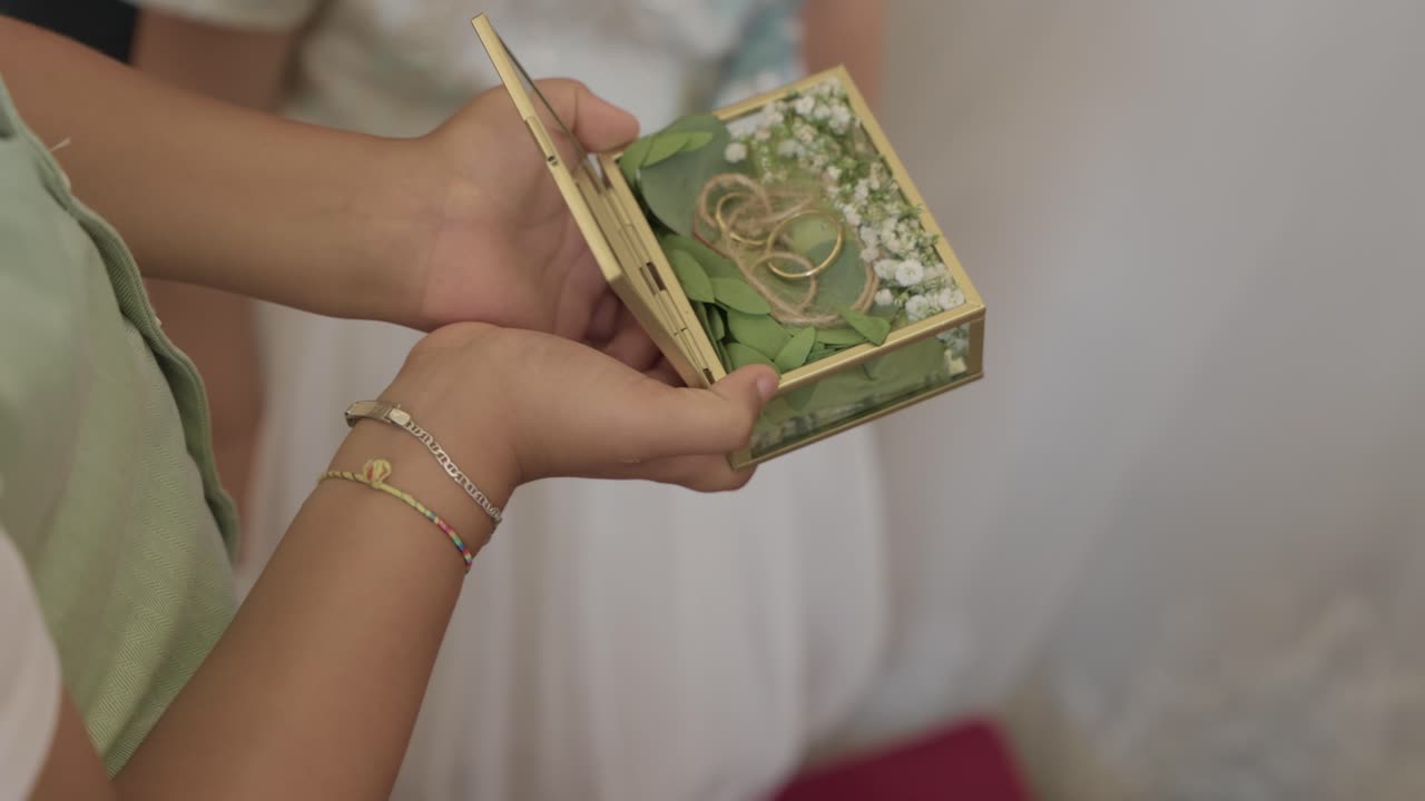 초록 잎과 흰 꽃으로 장식된 작은 유리 상자 안에 한 쌍의 결혼반지가 놓여 사랑과 결합을 상징하고 있어요.