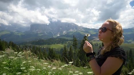 blonde girl blowing a dandelion flower in the wind, beautiful mountains nature setting sunny summer day at seiser alm, val gardena, dolomiti