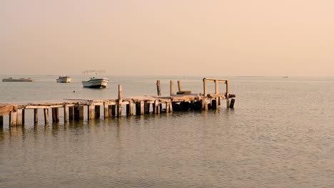 Antiguo-Muelle-De-Madera-Sobre-Aguas-Tranquilas-Del-Mar-Sobre-El-Cielo-Del-Atardecer-Con-Barcos-En-El-Fondo,-Bahrein