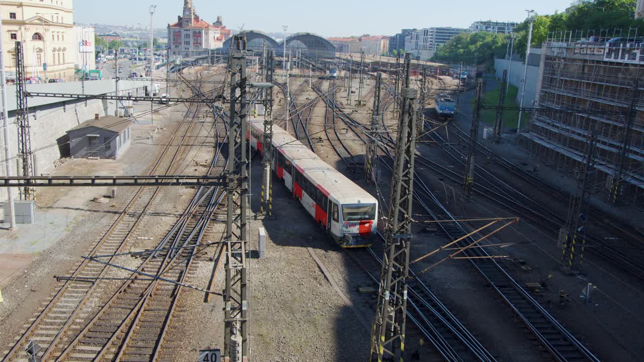 A red and white passenger train approaches the main railway station in Prague under bright daylight, captured from an elevated, static viewpoint