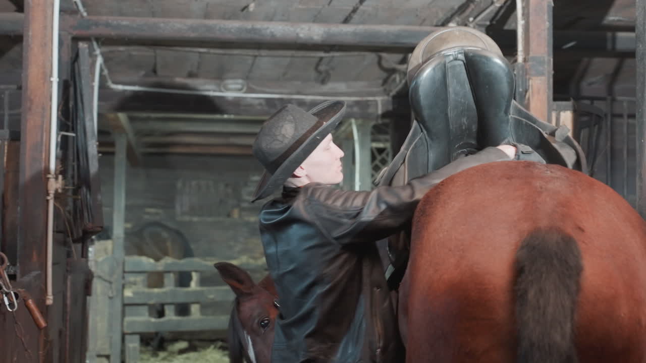 Uomo bianco che sistema la sella su un cavallo in una stalla, con l'istruttore che dimostra la regolazione del sottopancia e i controlli di equilibrio. Attrezzatura in cuoio e cappello da cowboy, mani che fissano l'equipaggiamento, educazione pratica e guida in tandem.