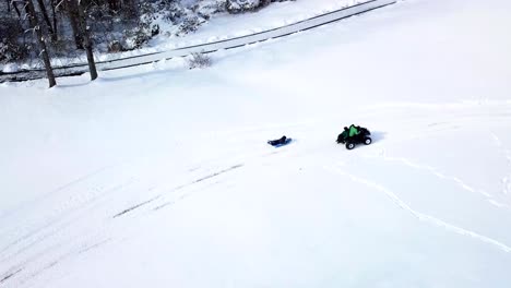 a drone, aerial, birds eye top down view of a child riding on their belly on a sled getting pulled by an atv 4 wheeler with a child sitting on the back in the snow covered ground of the country