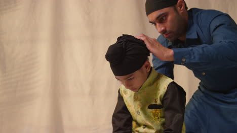studio shot of father tying turban onto head of young sikh son wearing traditional waistcoat as sequence part 9 of 10