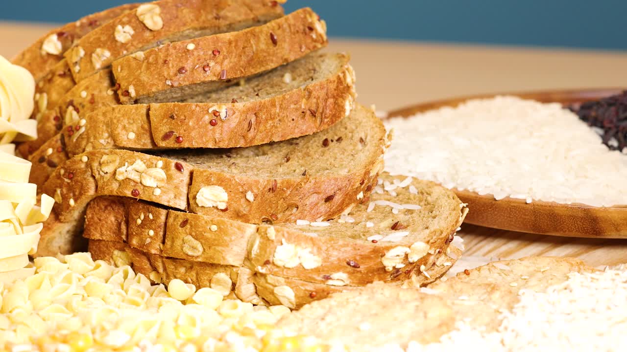 A variety of grains and sliced bread are artfully arranged on a wooden surface under bright lighting, showcasing texture and color