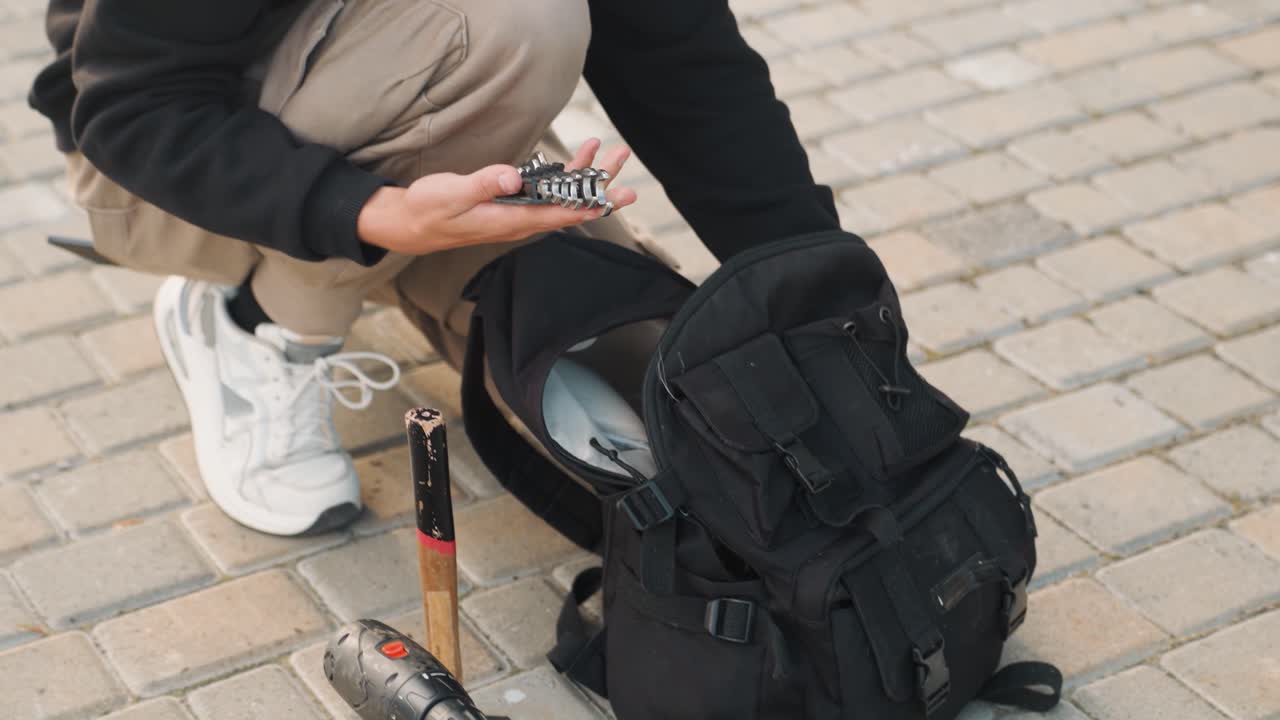 Mladý bílý student skrčený vedle batohu, vytahující nástroje ze sady na chodníku, soustředěné ruce upravující hardware skútru, ležérní mikina s kapucí a cargo kalhoty, bílé tenisky, městský chodník za denního světla.