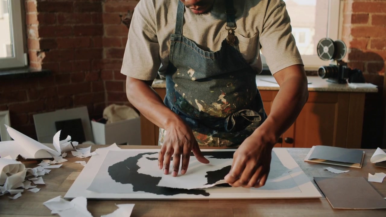 Lifting torn sheet, apron-clad artist reshaping on studio table, placing and assembling silhouette