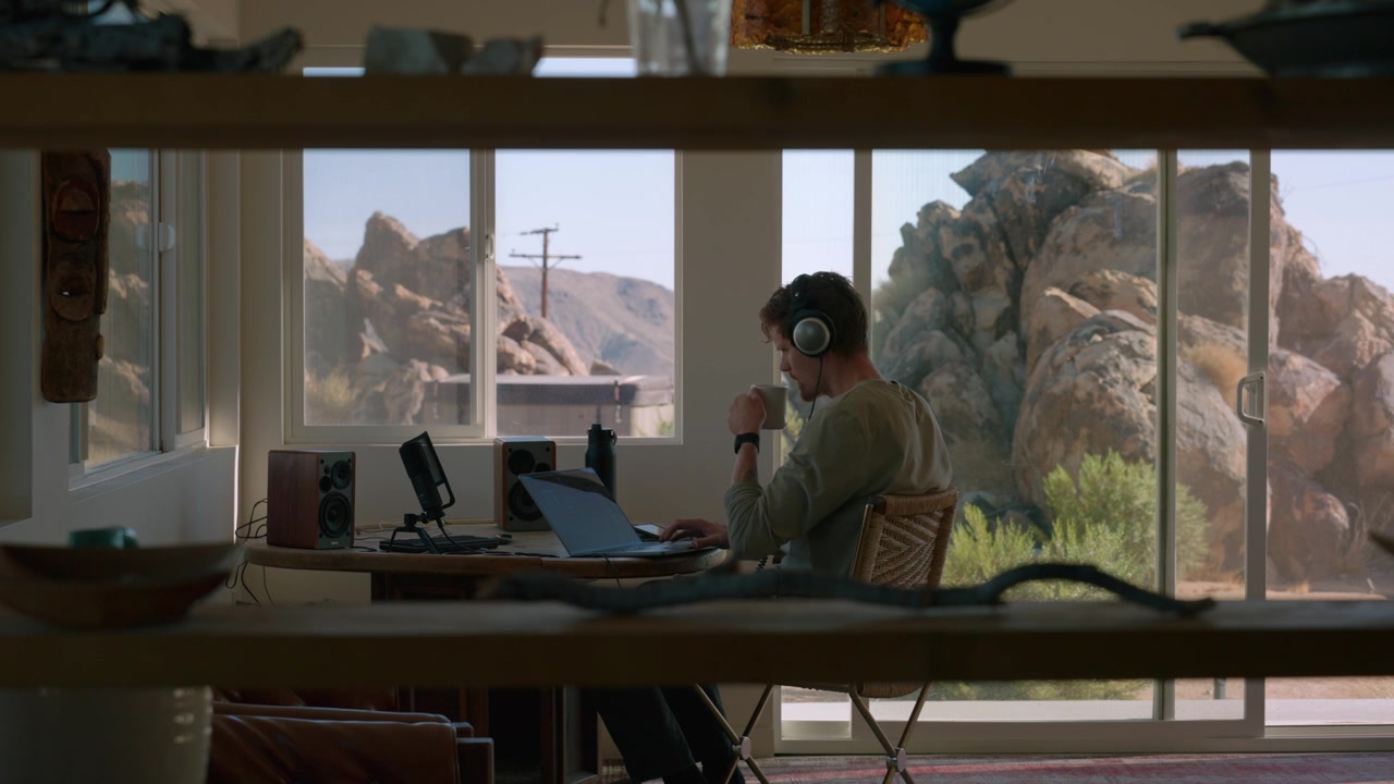 Wearing headphones, a man works at a desk with a laptop and coffee overlooking the desert.