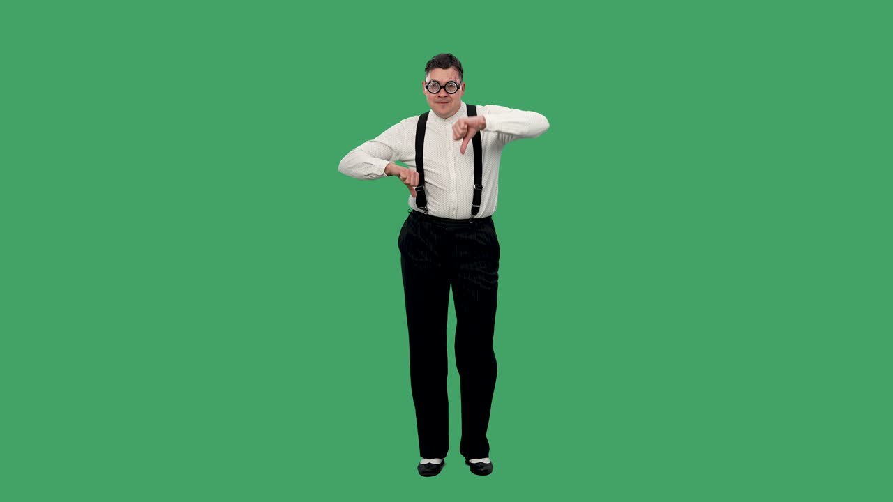 춤을 추고 두 손으로 엄지손가락을 보여주는 남자의 초상화: 두꺼운 렌즈를 입은 안경,  셔츠와 서펜더를 입은 남자가 녹색 스크린에서 스튜디오에서 포즈를 취합니다.