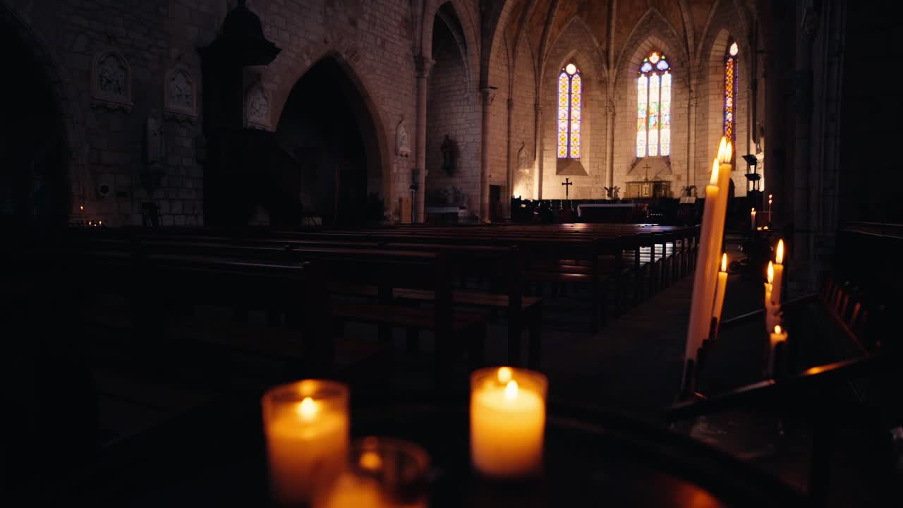 Række af brændende stearinlys i et mørkt kirkeinteriør i Monpazier, Dordogne, med træbænke og gotisk farvet glas. Slowmotion sporingsoptagelse
