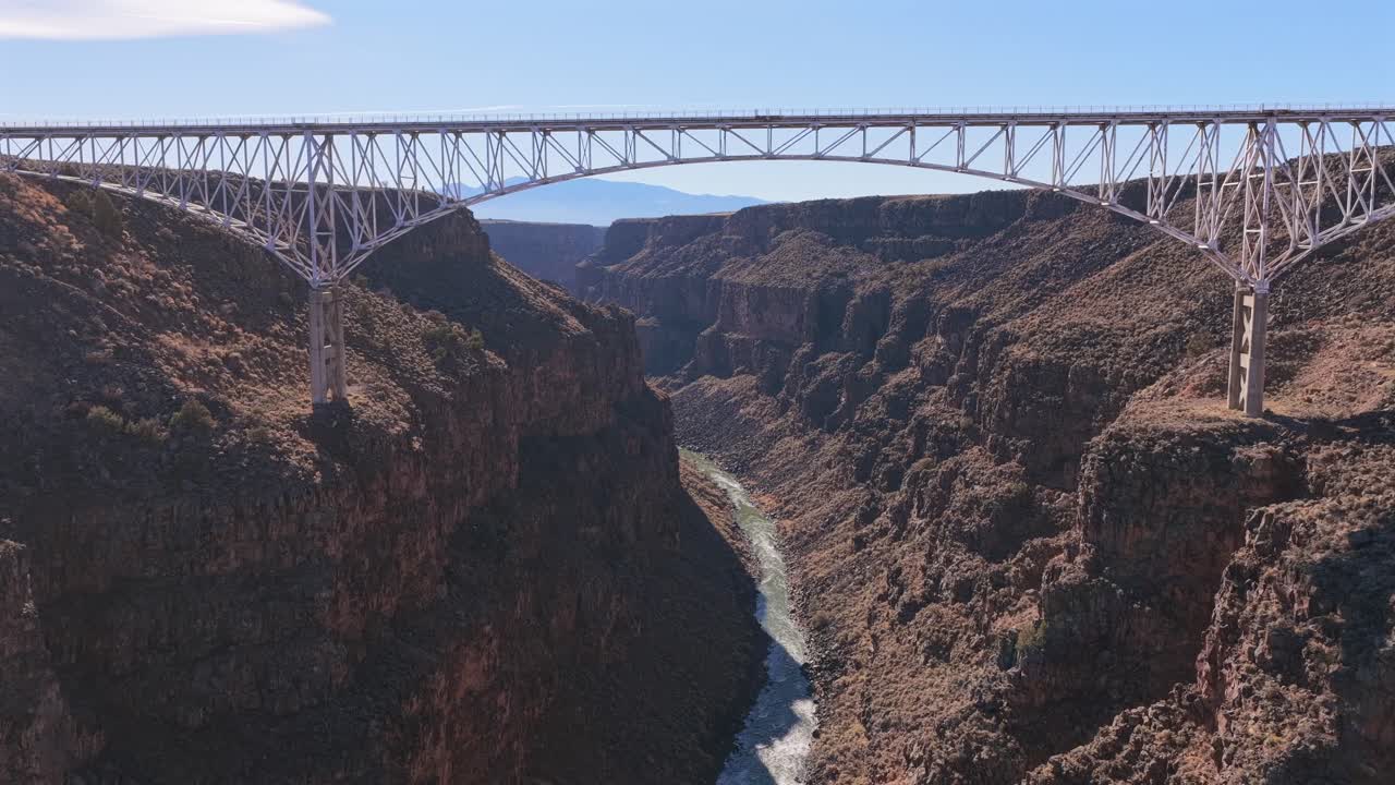 A wide aerial view shows a car driving across the steel Rio Grande Gorge Bridge. The bridge spans a deep rocky canyon in the high desert of New Mexico under a clear, bright blue sky