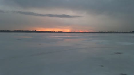 Vista-Aérea-De-Un-Lago-Congelado-Con-Una-Colorida-Puesta-De-Sol-En-El-Horizonte-En-Un-Día-Nublado