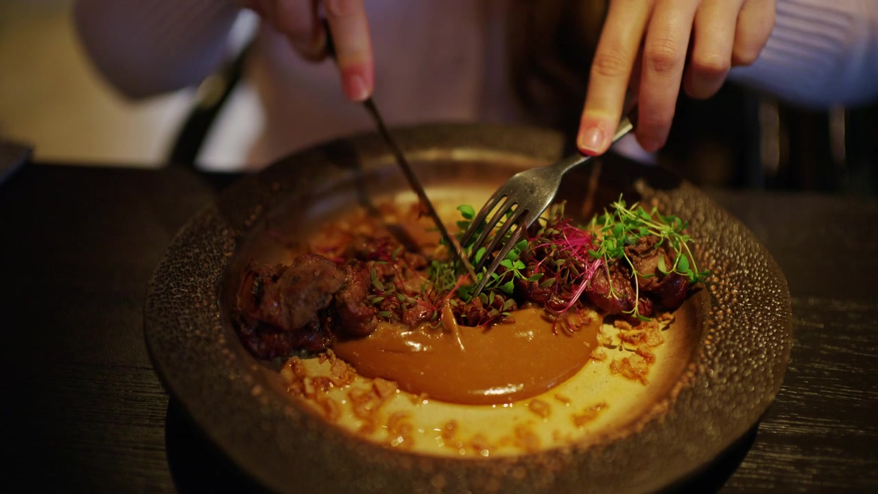 Close up of a gourmet fine dining dish being cut with fork and knife. Elegant plating with tender meat, creamy sauce, and fresh microgreens in a stylish restaurant setting