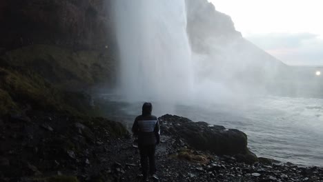 Girl-standing-under-waterfall-slowmotion-shot-from-bottom-to-top-in-Iceland-dark-cave