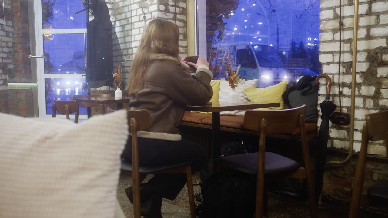 Back shot of female in brown shearling jacket sitting at cozy cafe sipping coffee, bus passing outside window creating a lively and peaceful moment in the background