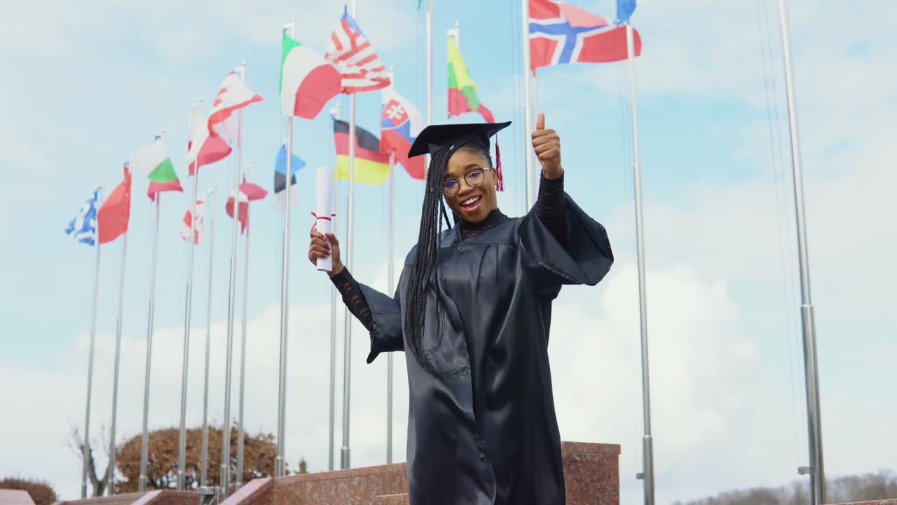 una joven afroamericana se alegra con un diploma de educación superior en la mano en la calle contra el fondo de banderas de diferentes países de europa y el mundo